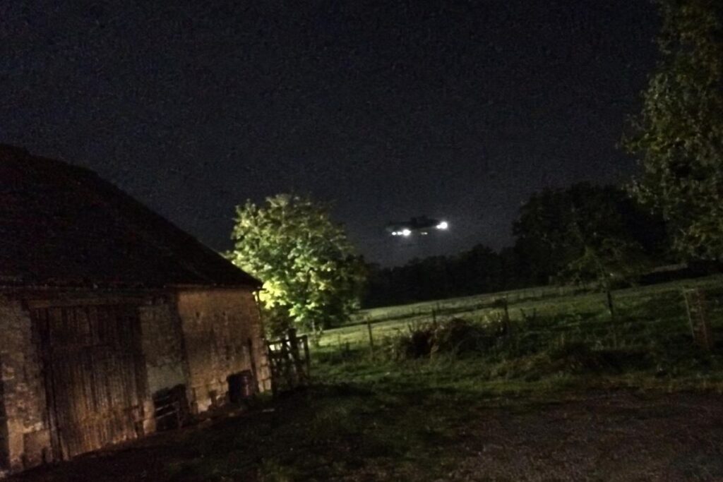 Forme sombre et lumières blanches en vol stationnaire au-dessus d’un champ près d’une grange