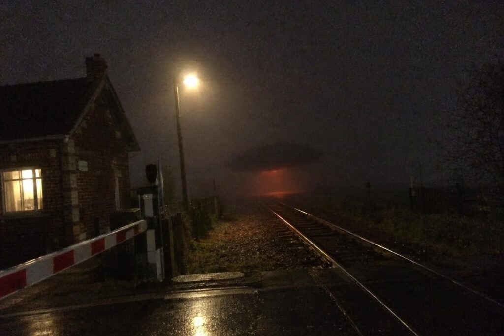 Illustration : masse sombre éclairée de rouge au-dessus d’une voie ferrée, près d’un passage à niveau, de nuit dans le brouillard