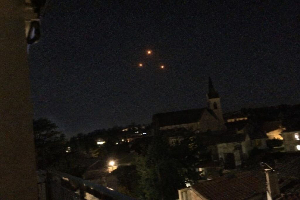 Illustration : trois lumières orange en triangle dans le ciel nocturne au-dessus d’un village