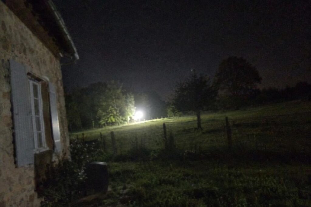 Illustration : forte lumière blanche au ras d’un champ derrière une ferme, de nuit