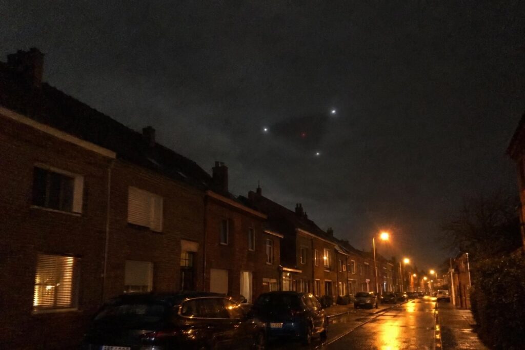 Illustration : triangle sombre à trois lumières blanches et lumière rouge au-dessus d’une rue belge, de nuit